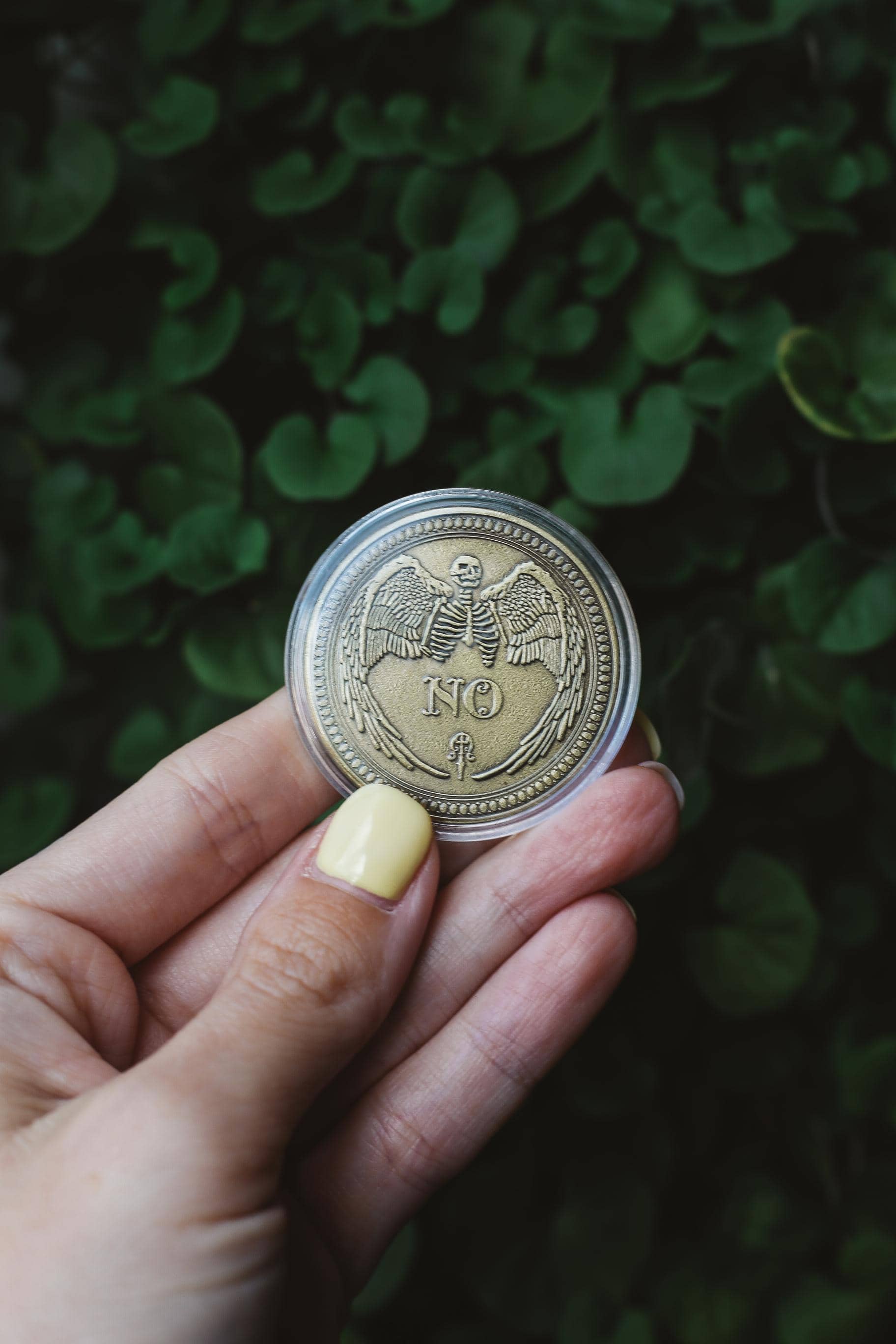 Hand holding a fortune coin featuring winged skeleton with a blurred green leafy background