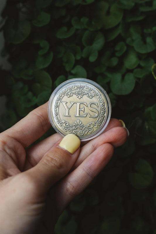 Hand holding a coin with 'YES' engraved on it against a green leafy background