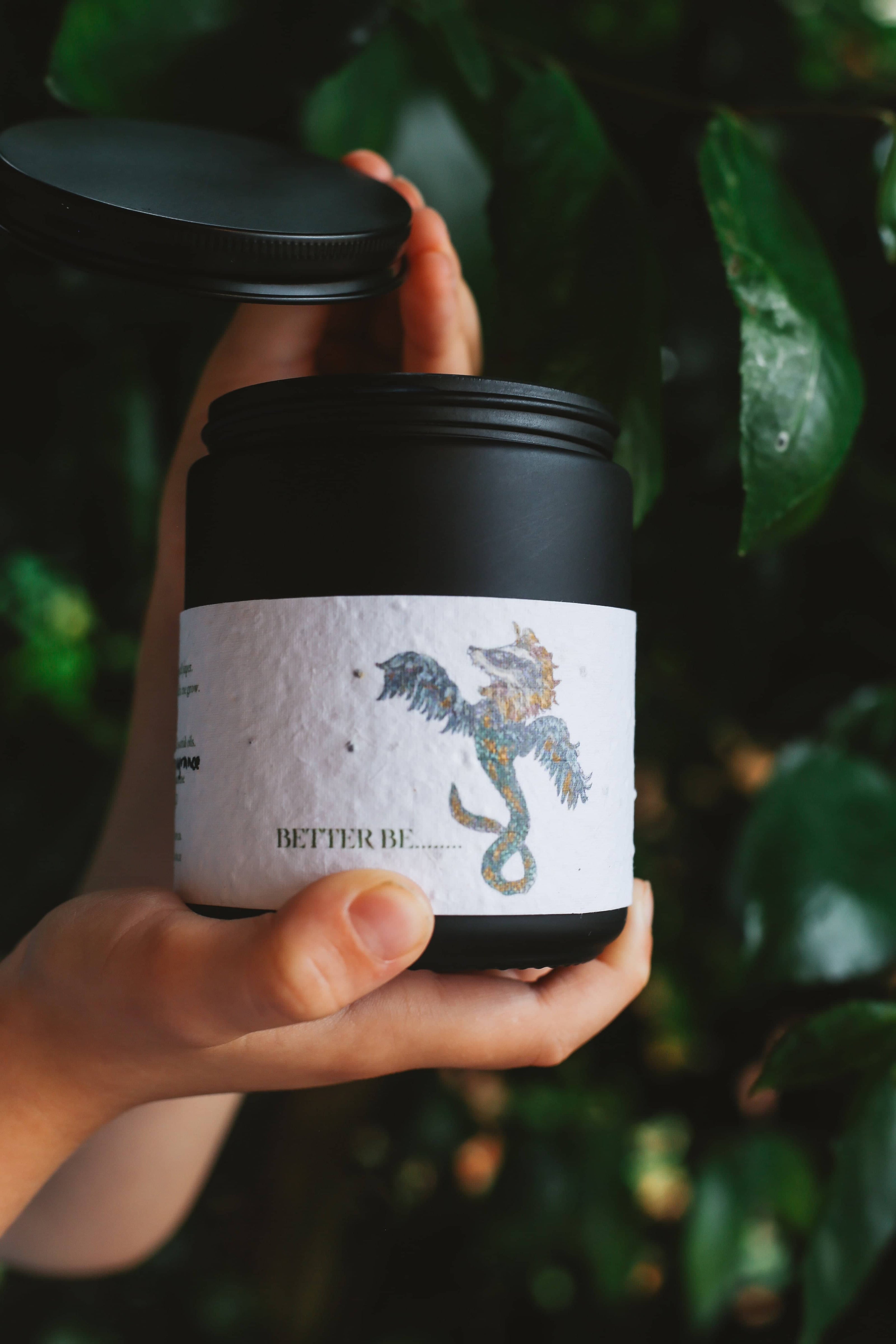 Hand holding Àine & Co. "Better Be..." Revealing Candle, packaged in matte black jar with matte black aluminium lid, with seed paper label