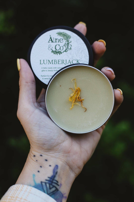 Hand holding a tin of 'Lumberjack' balm with 'Aine & Co' branding against a dark background.