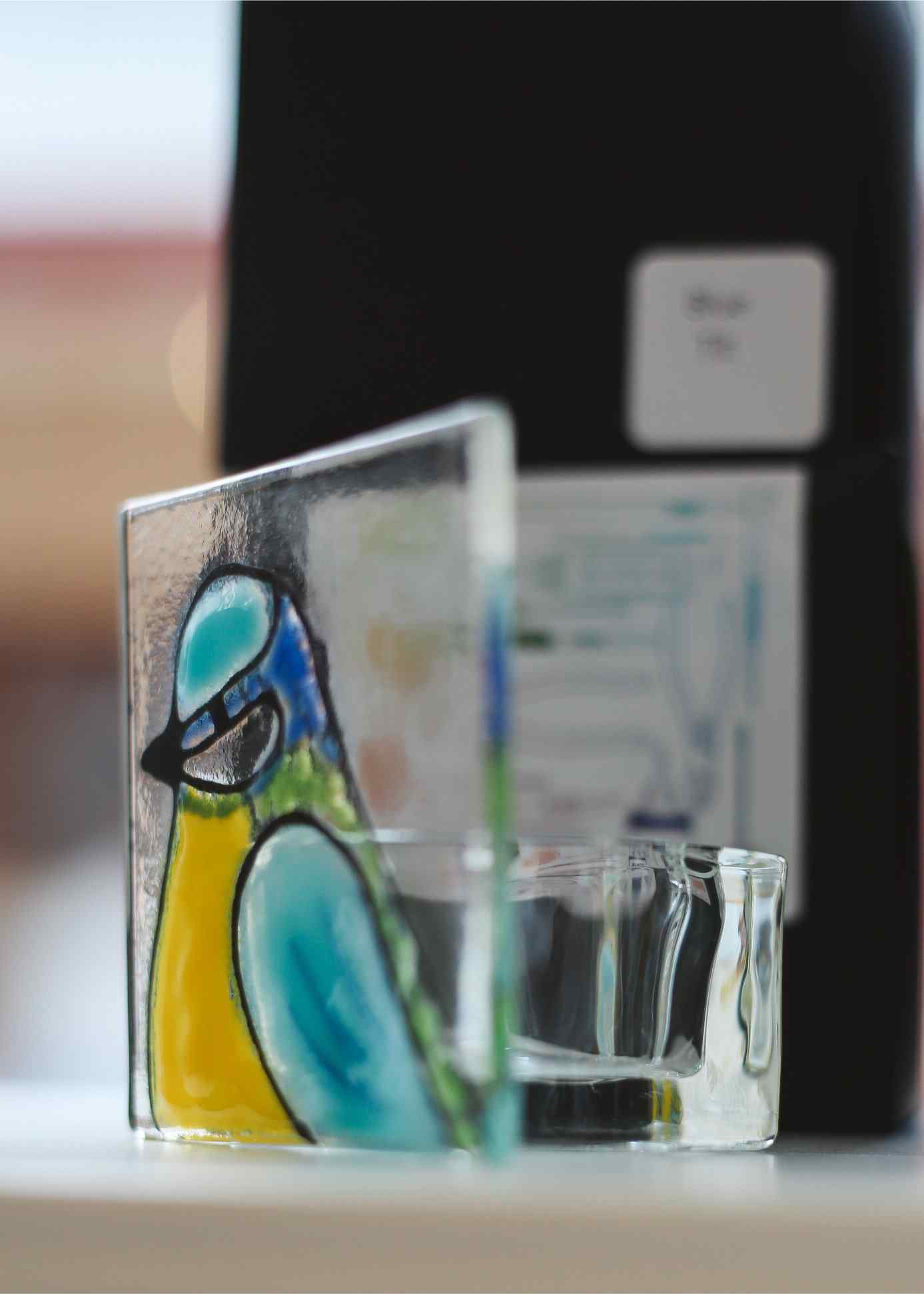A clear glass candle holder with a colorful painted bird design in blue, yellow, and green, shown on a tabletop with a blurred dark bottle in the background.