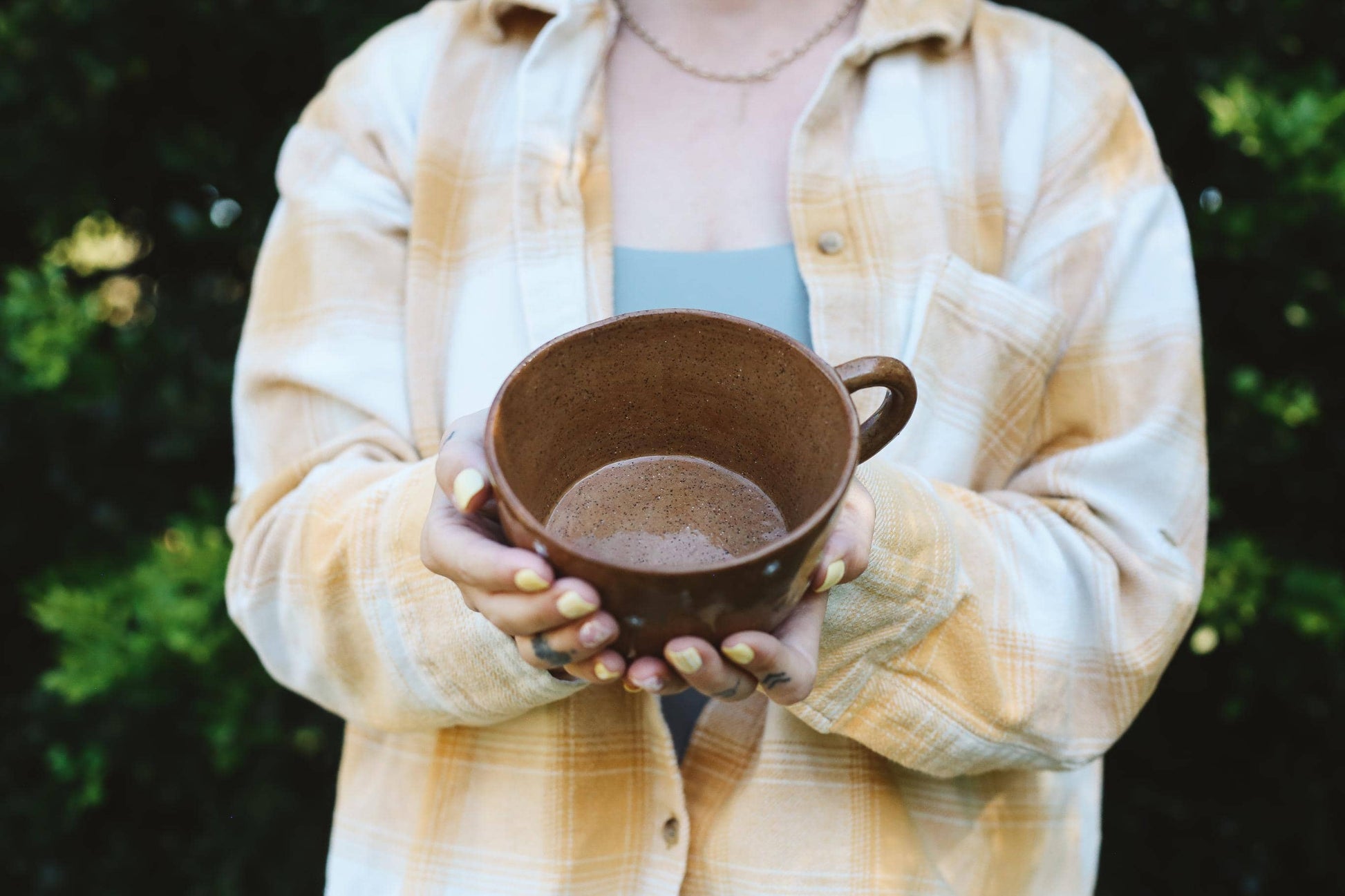 Person holding a brown ceramic cup outdoors with greenery in the background