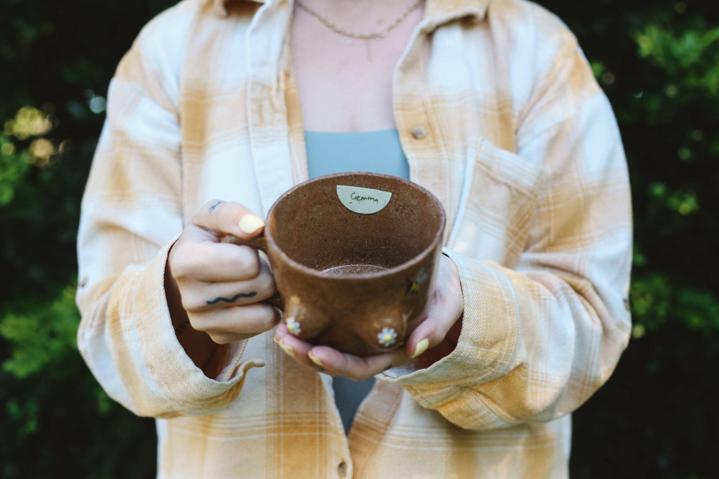 Person holding a brown ceramic cup outdoors with greenery in the background