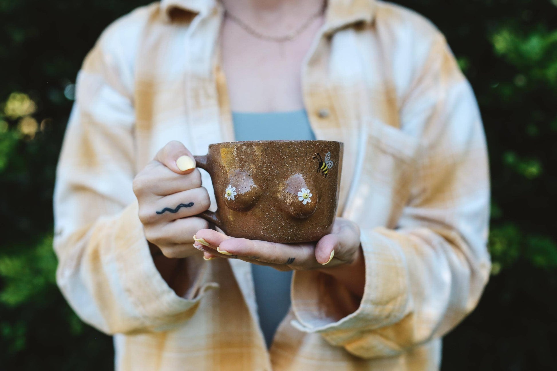 Person holding a brown ceramic mug with bee and daisy designs outdoors