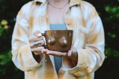 Person holding a brown ceramic mug with bee and daisy designs outdoors