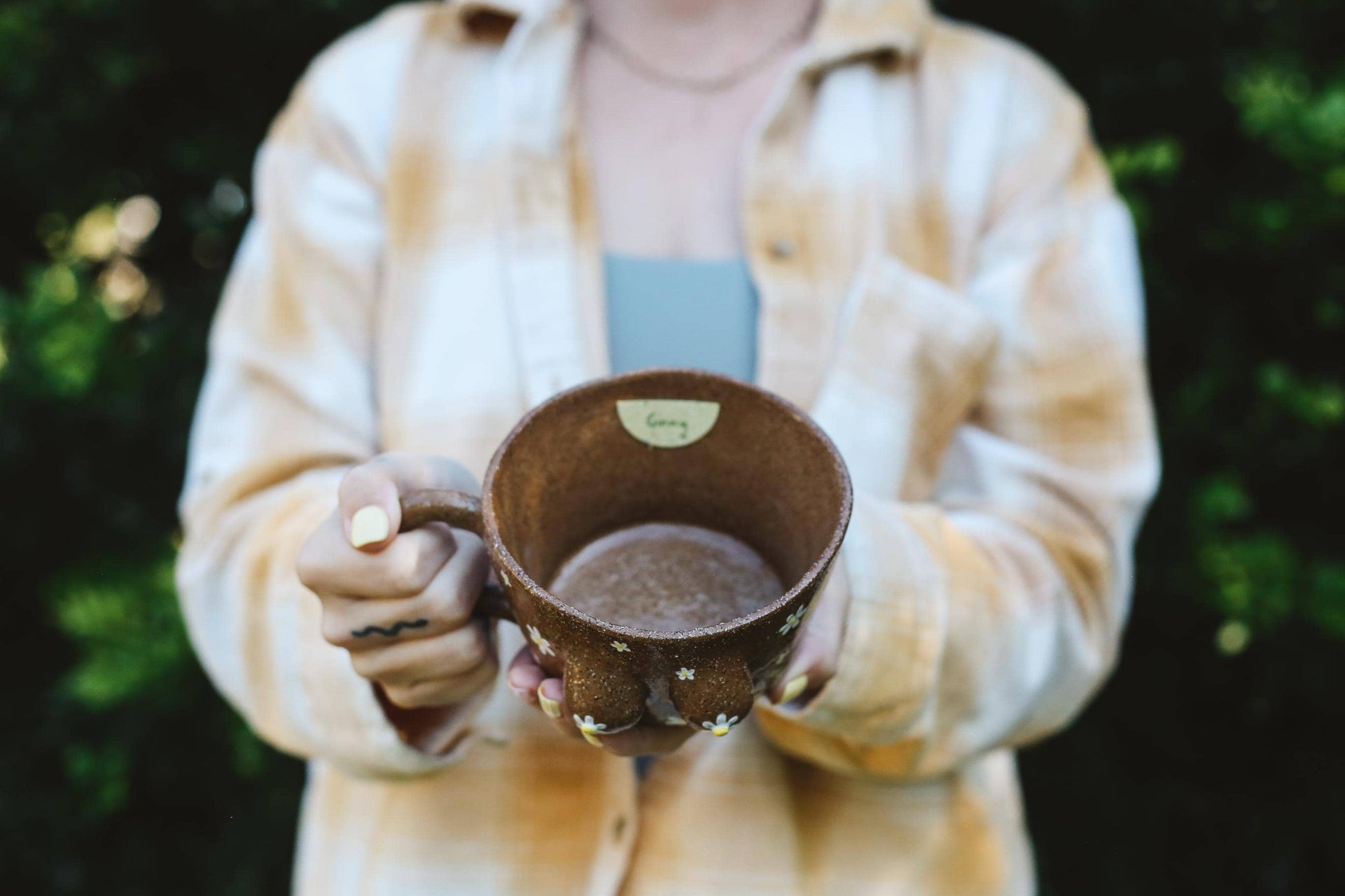 Person holding a ceramic mug outdoors with a blurred green background