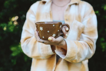 Person holding a brown mug with white floral patterns against a blurred green background