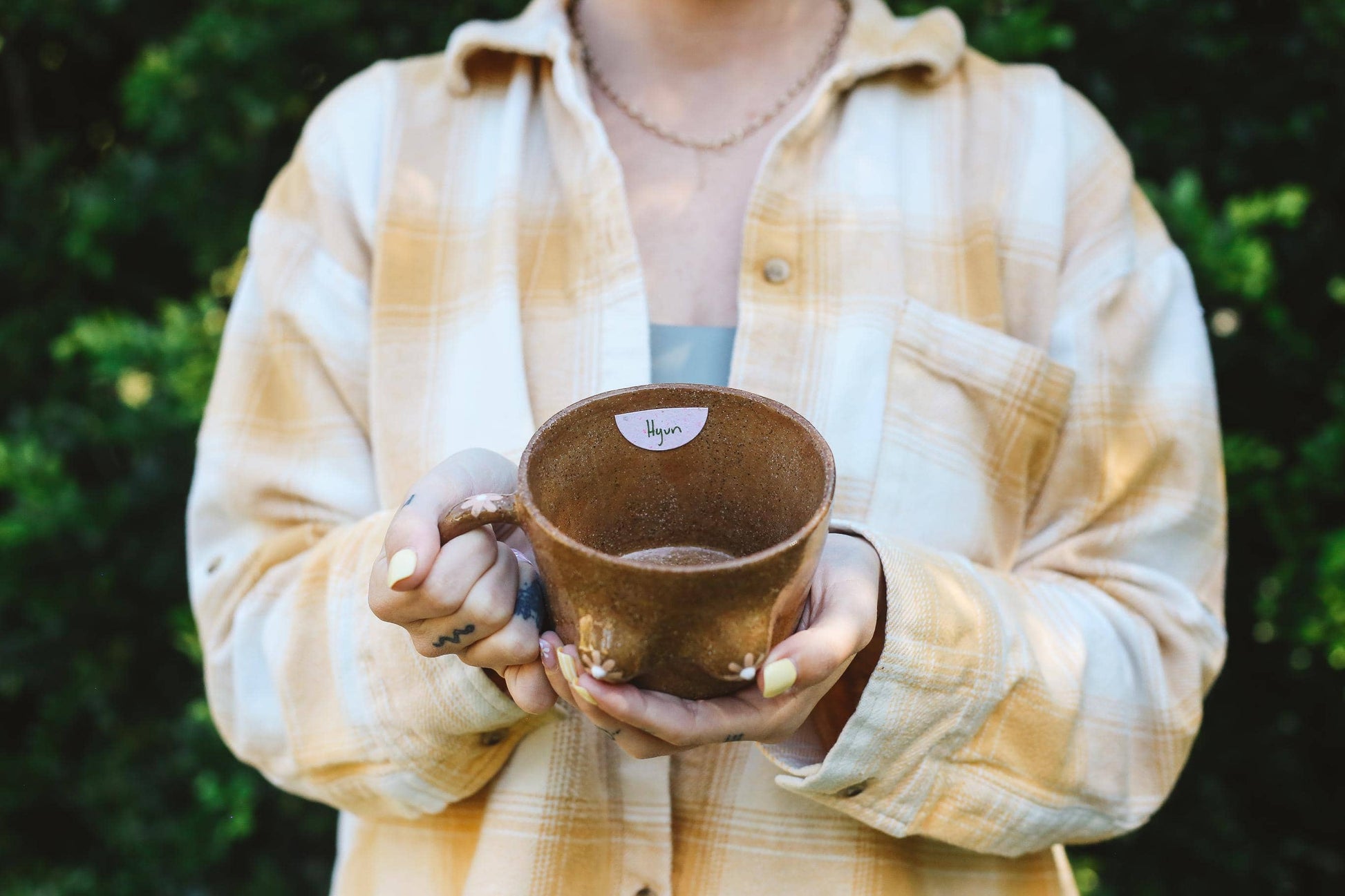 Person holding a brown ceramic mug outdoors with greenery in the background