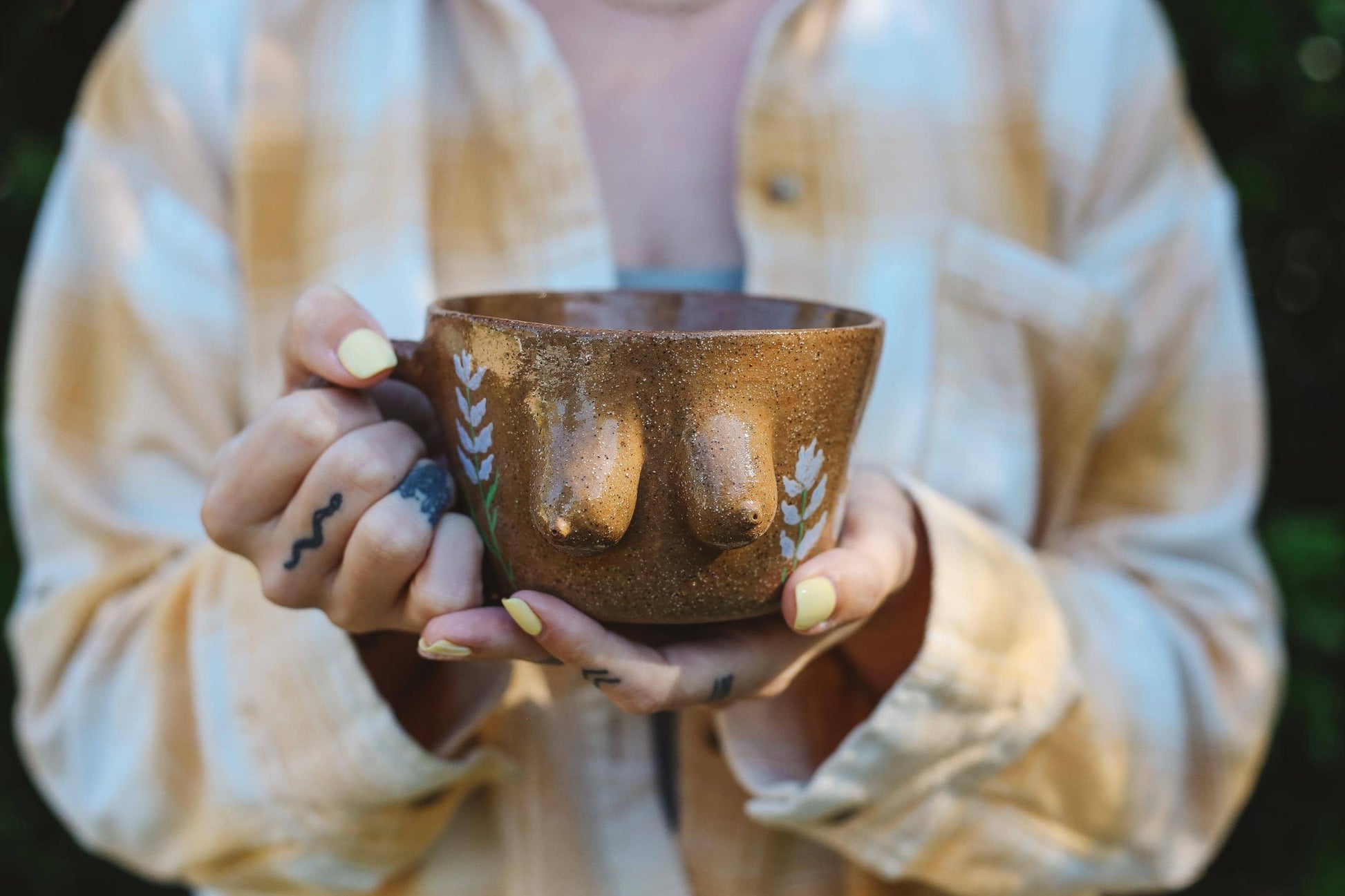 Person holding a brown ceramic mug with a breast design against a blurred background
