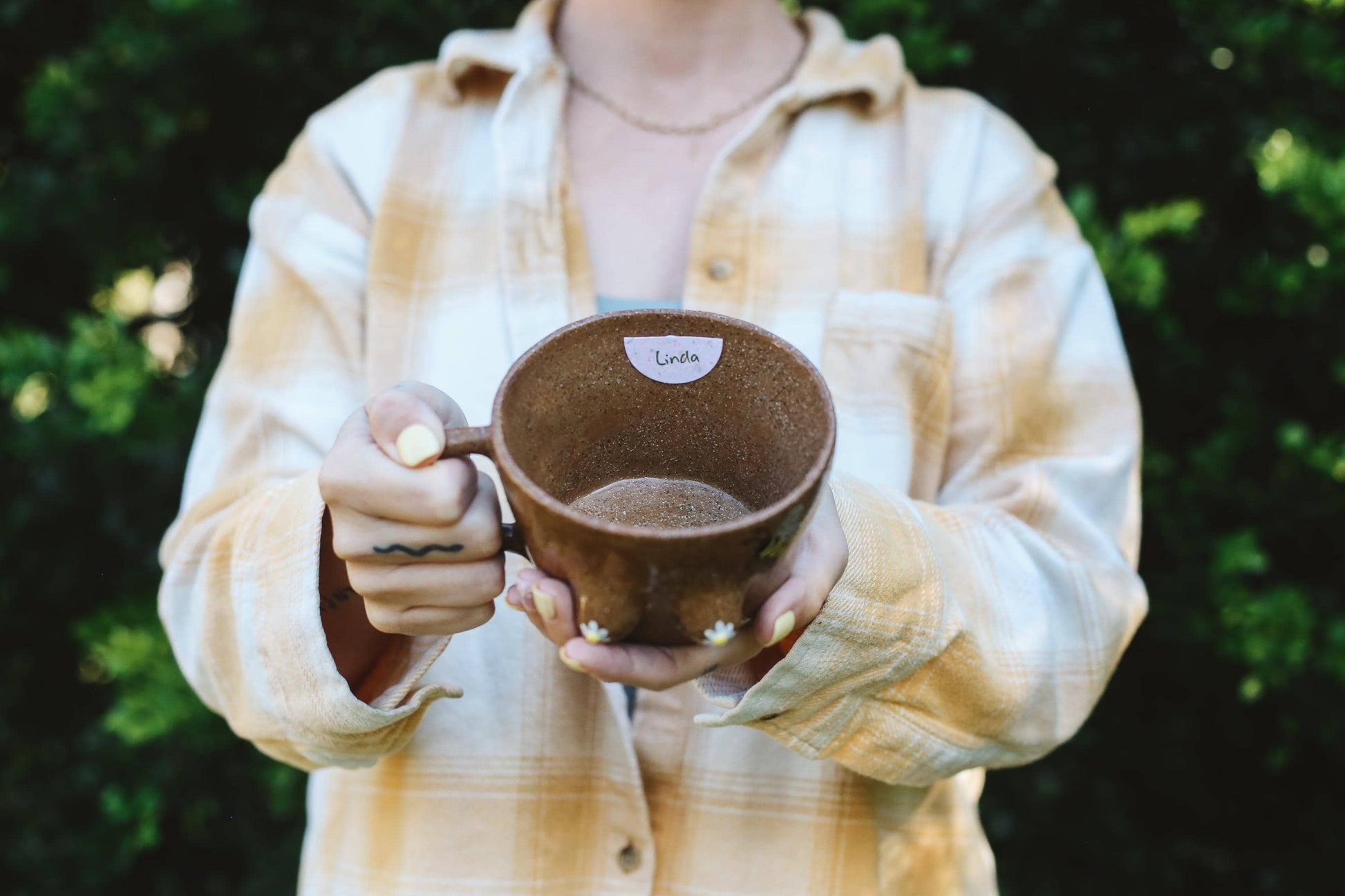 Person holding a brown mug outdoors with greenery in the background