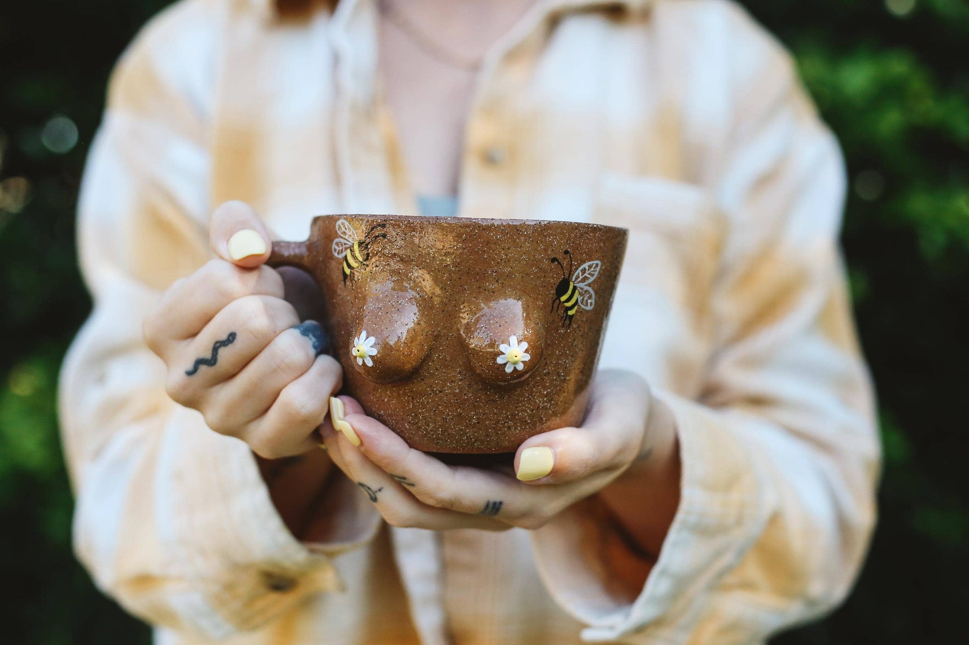 Person holding a brown mug with a boob and bumblebee design against a blurred green background