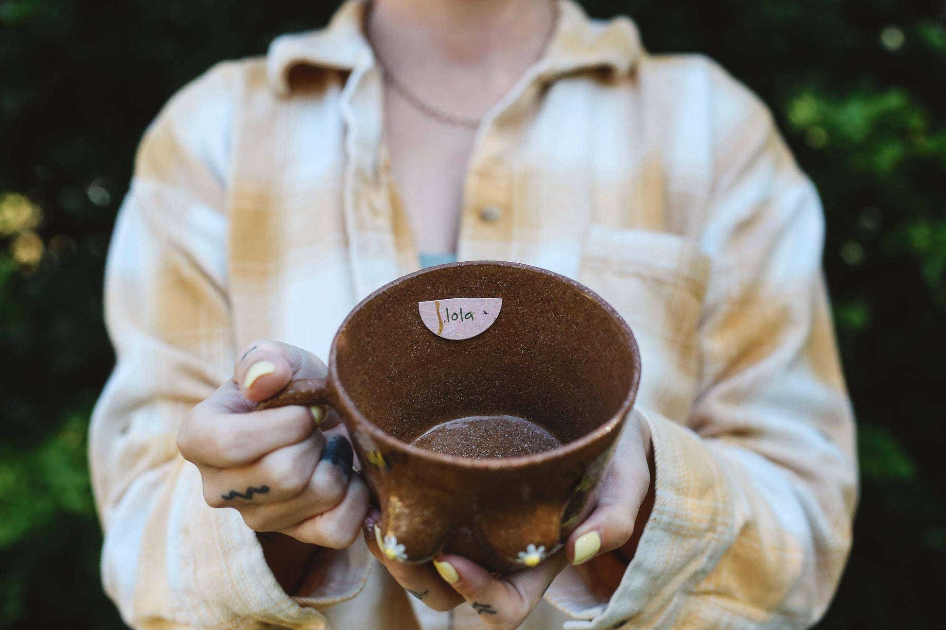 Person holding a brown ceramic mug with name tag "Lola" against a blurred natural background