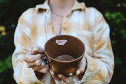 Person holding a brown ceramic mug with name tag "Lola" against a blurred natural background