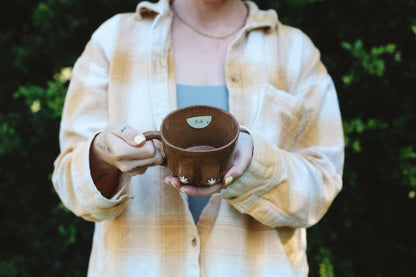 Person holding a brown ceramic mug outdoors with greenery in the background