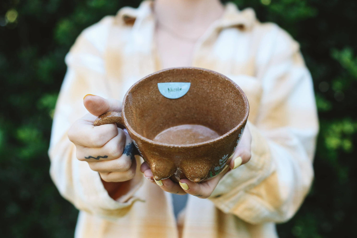 Person holding a brown ceramic cup outdoors with greenery in the background