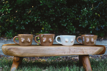 Four ceramic mugs on a wooden stool outdoors with greenery in the background