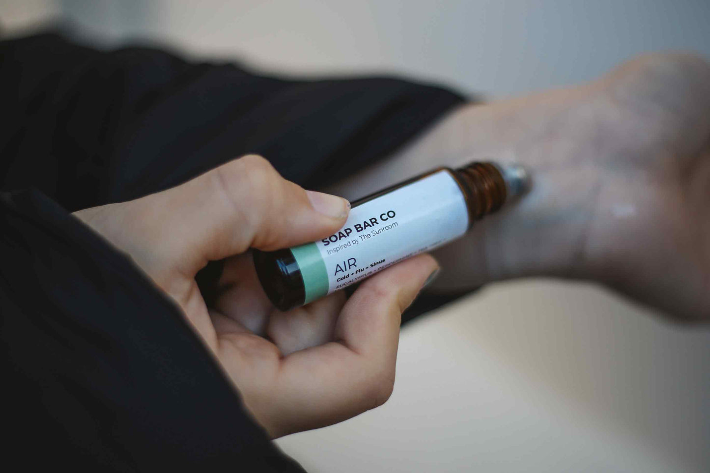 A close-up of a person holding a Soap Bar Co. rollerball bottle over their foot, applying a clear gel treatment.