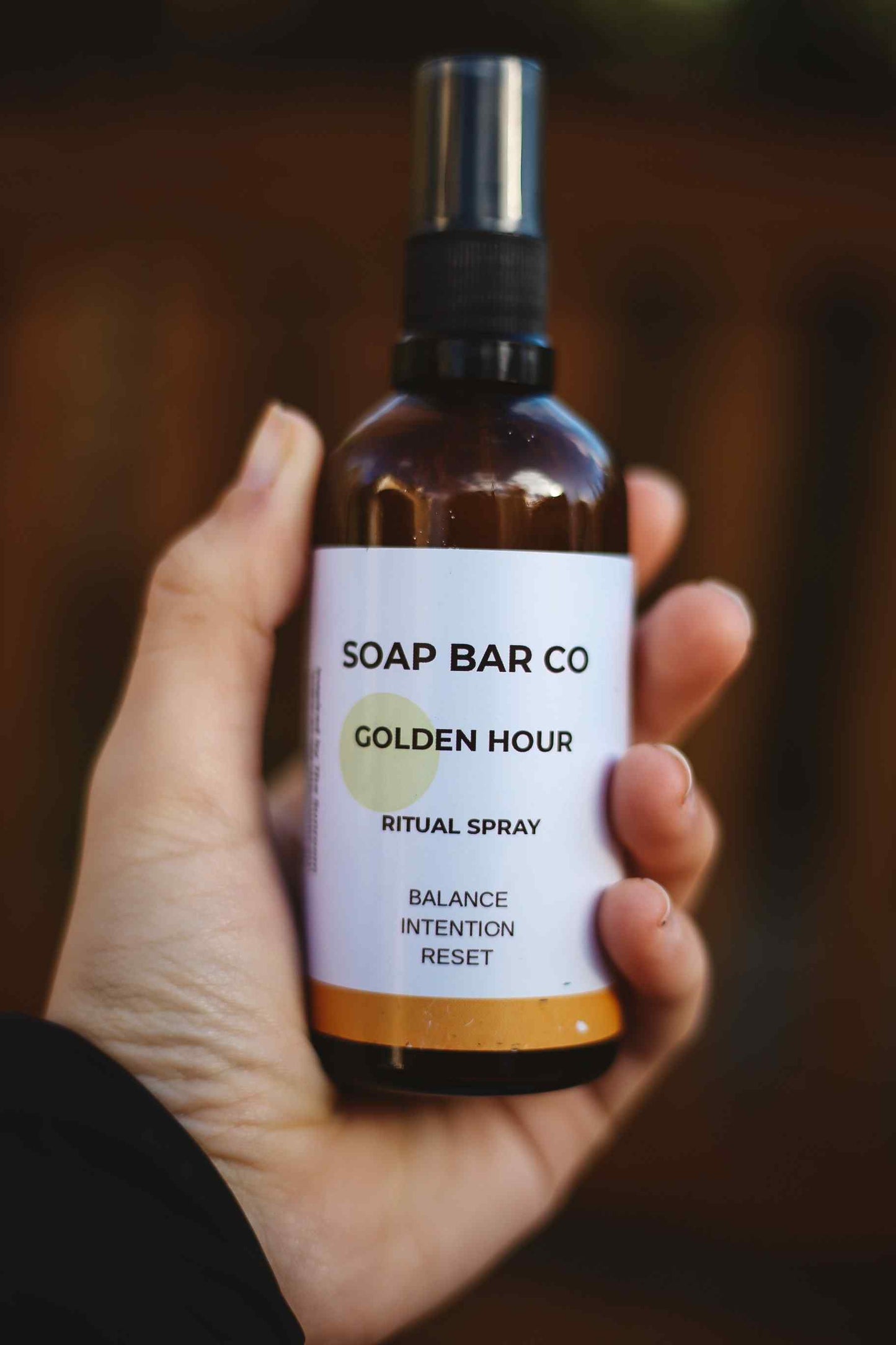 A hand holding an amber glass spray bottle labeled 'SOAP BAR CO GOLDEN HOUR RITUAL SPRAY' against a dark blurred background.