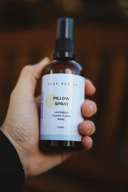 A hand holding a 100ml amber glass spray bottle of Soap Bar Co Pillow Spray (Lavender, Ylang Ylang, Rose) against a dark, blurred background.