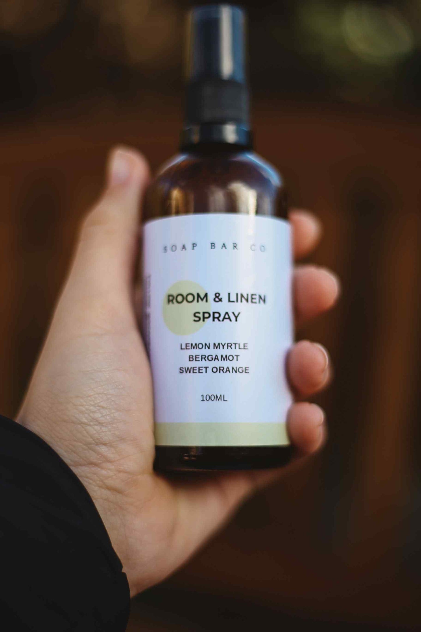 A hand holding a 100ml spray bottle of Soap Bar Co. Room & Linen Spray in Lemon Myrtle Bergamot and Sweet Orange, set against a dark, blurred background.