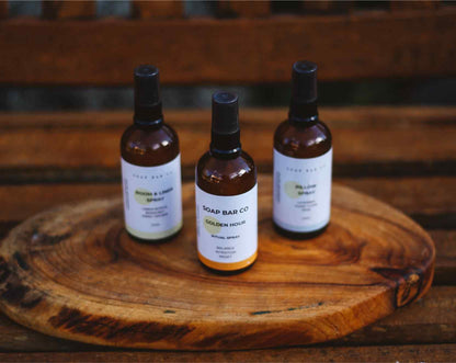 Three amber glass spray bottles with black caps and white labels from Soap Bad Co. are arranged on a round wooden serving tray, set against a rustic wooden background.