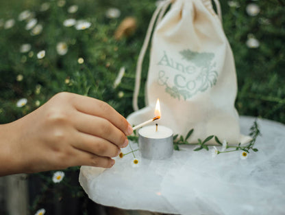 A hand lighting an Àine & Co. Tea Light with an Àine & Co. Perfect Match, shown with the Àine & Co. calico drawstring bag