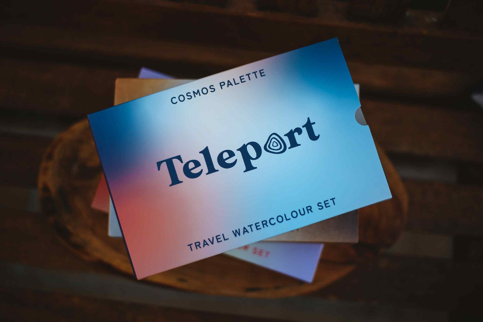 Teleport Cosmos Palette travel watercolour set booklet resting on a wooden surface, showing a blue-to-pink gradient cover with the Teleport logo.