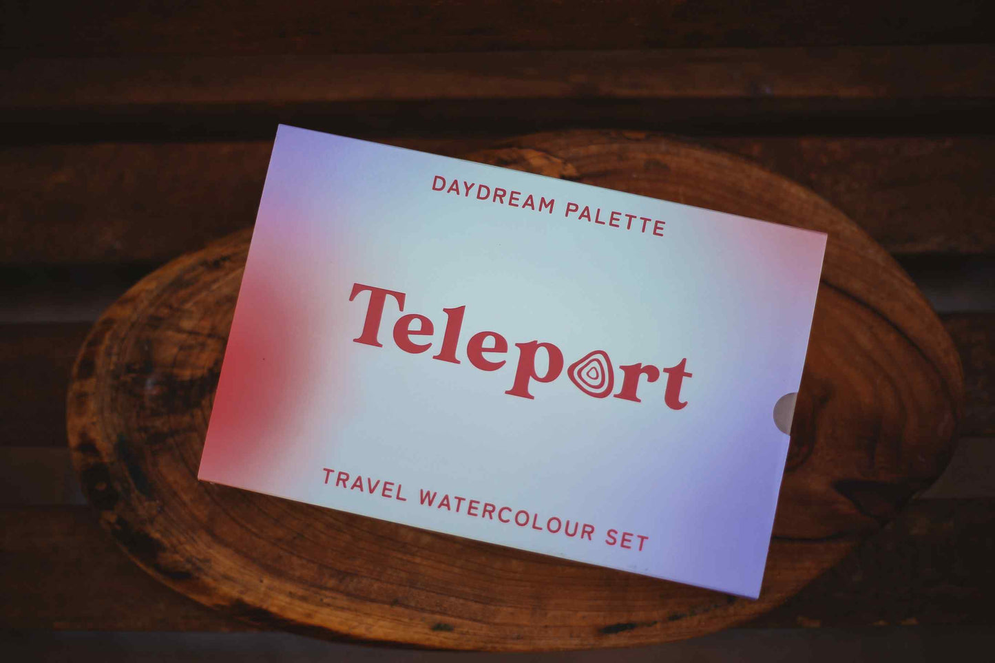 A Teleport Daydream Palette travel watercolour set card resting on a rustic wooden slice, with a dark wood background.
