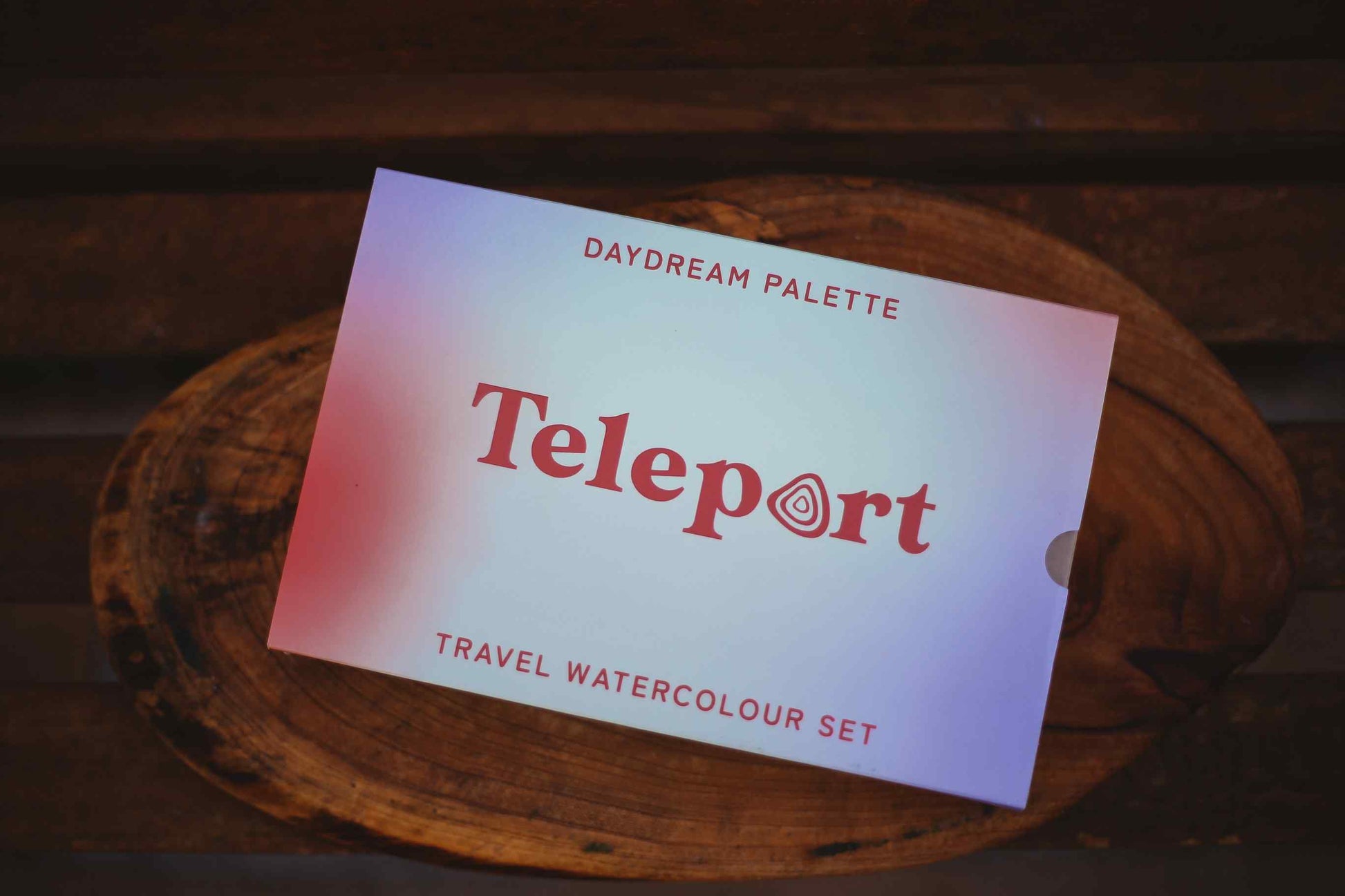 A Teleport Daydream Palette travel watercolour set card resting on a rustic wooden slice, with a dark wood background.