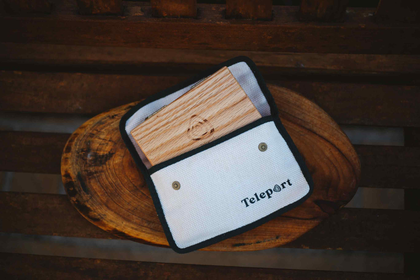 A white fabric pouch with black trim and the brand name 'Teleport' printed on it, holding a rectangular wooden block, displayed on a rustic wooden slice over a dark wooden surface.