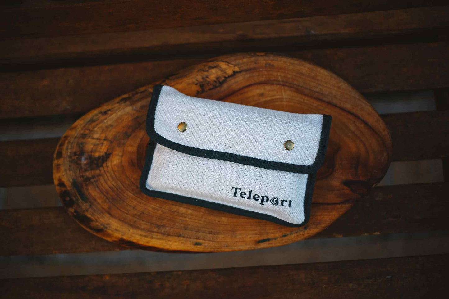 A white fabric pouch with black trim and the word 'Teleport' printed on the front, resting on a rustic wooden slice over a dark wooden surface.