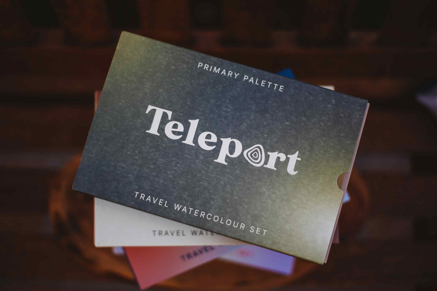 Teleport Primary Palette travel watercolour set, showing the dark textured cover with white text, resting on a stack of other color palettes.