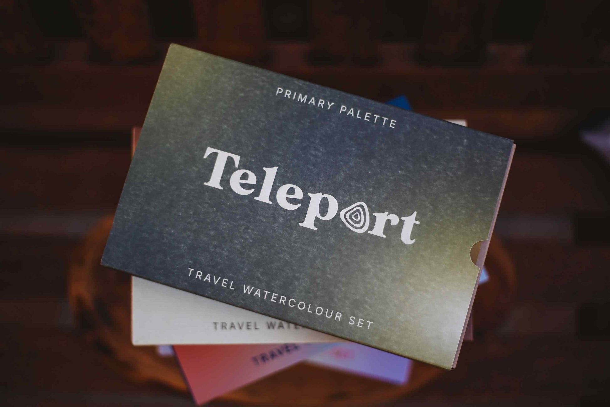 Teleport Primary Palette travel watercolour set, showing the dark textured cover with white text, resting on a stack of other color palettes.