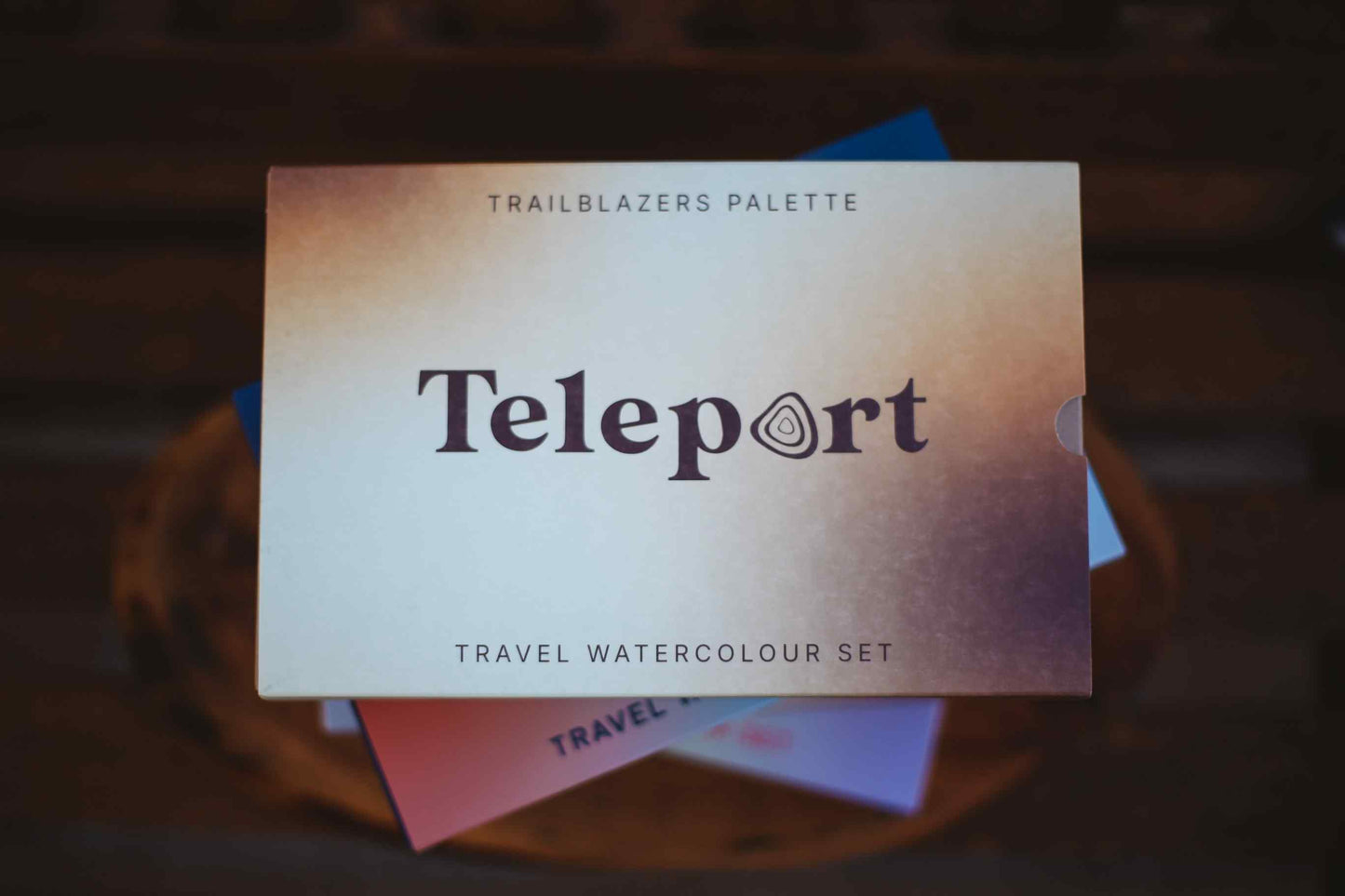 Teleport Trailblazers Palette travel watercolour set, featuring a square palette cover with a warm beige-to-purple gradient and the word 'Teleport' in dark purple text, resting on a dark wooden surface.