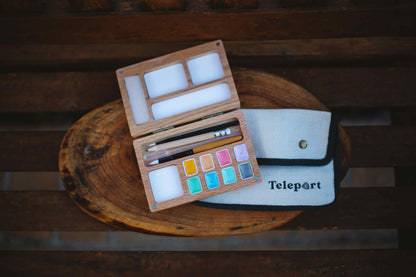 An open wooden watercolor paint palette with white blocks, paintbrushes, and a small canvas pouch labeled 'Teleport' on a rustic wooden slice background.